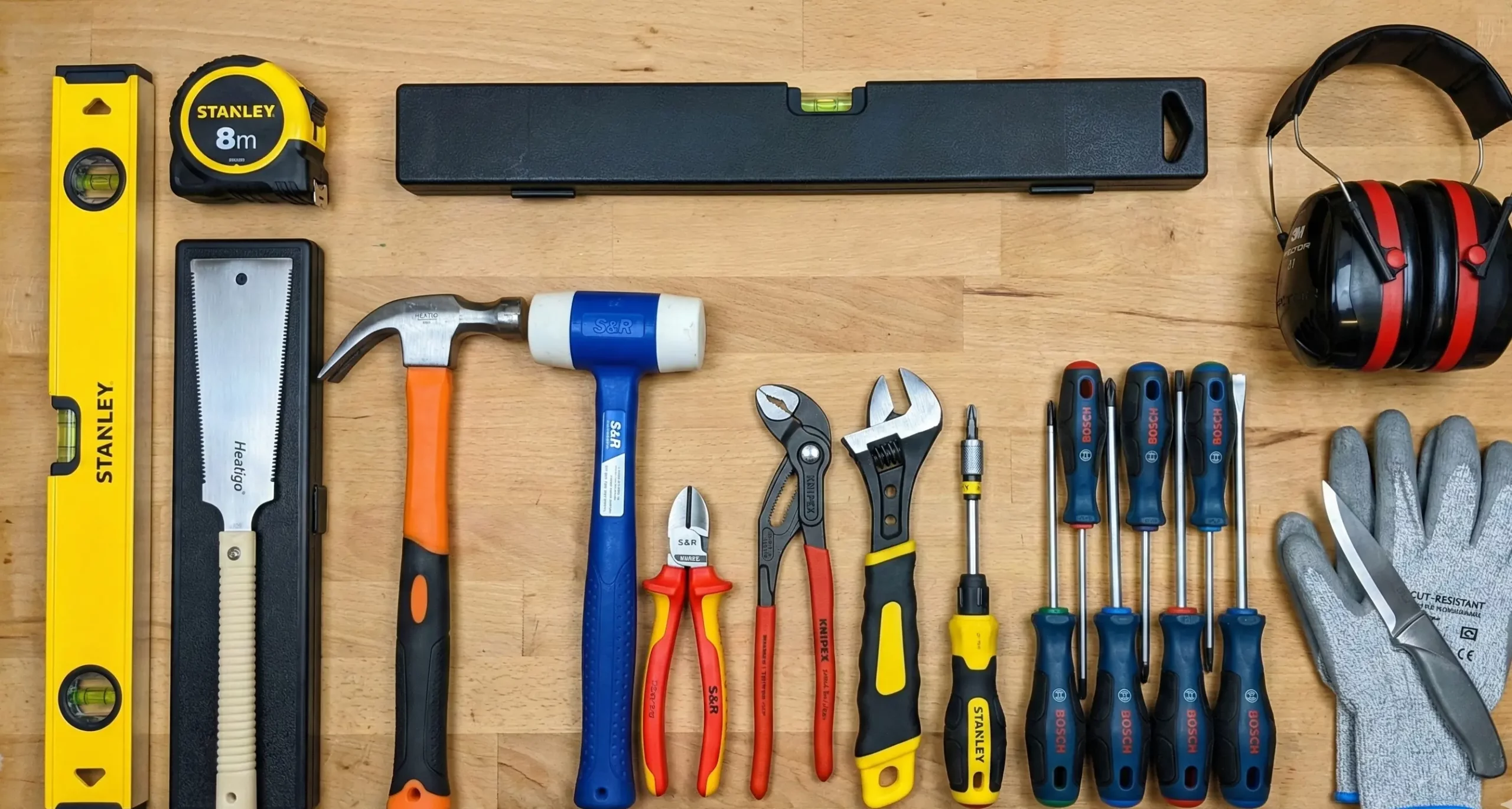 Photographie en vue de dessus montrant un assortiment complet d'outils manuels et d'équipements de protection individuelle soigneusement rangés sur une surface en bois clair. L'ensemble comprend, de gauche à droite : un niveau à bulle jaune Stanley, un mètre ruban Stanley de 8 mètres, une scie japonaise avec manche clair dans un étui noir, une longue boîte de rangement noire, un marteau arrache-clous à manche orange et noir, un maillet à tête blanche et manche bleu S&R, une pince coupante diagonale à manches rouges et jaunes S&R, une pince multiprise à manches rouges et noirs Knipex, une clé à molette à manche jaune et noir, un tournevis porte-embout Stanley, un jeu de six tournevis Bosch à manches bleus et rouges, un casque anti-bruit noir et rouge, ainsi qu'une paire de gants de travail gris sur lesquels est posé un cutter.