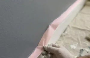 Gros plan d'une main gantée retirant doucement un ruban de masquage rose à 45 degrés d'un mur gris fraîchement peint.