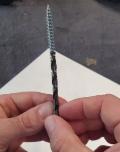 Astuce de menuisier pour savoir quel foret pour quelle vis choisir afin de pré-percer le bois sans le fendre. La photo montre en gros plan des mains d'artisan alignant une mèche à bois noire exactement derrière une vis argentée, prouvant visuellement que seul le filetage dépasse de chaque côté tandis que le corps central reste masqué par le diamètre du foret.